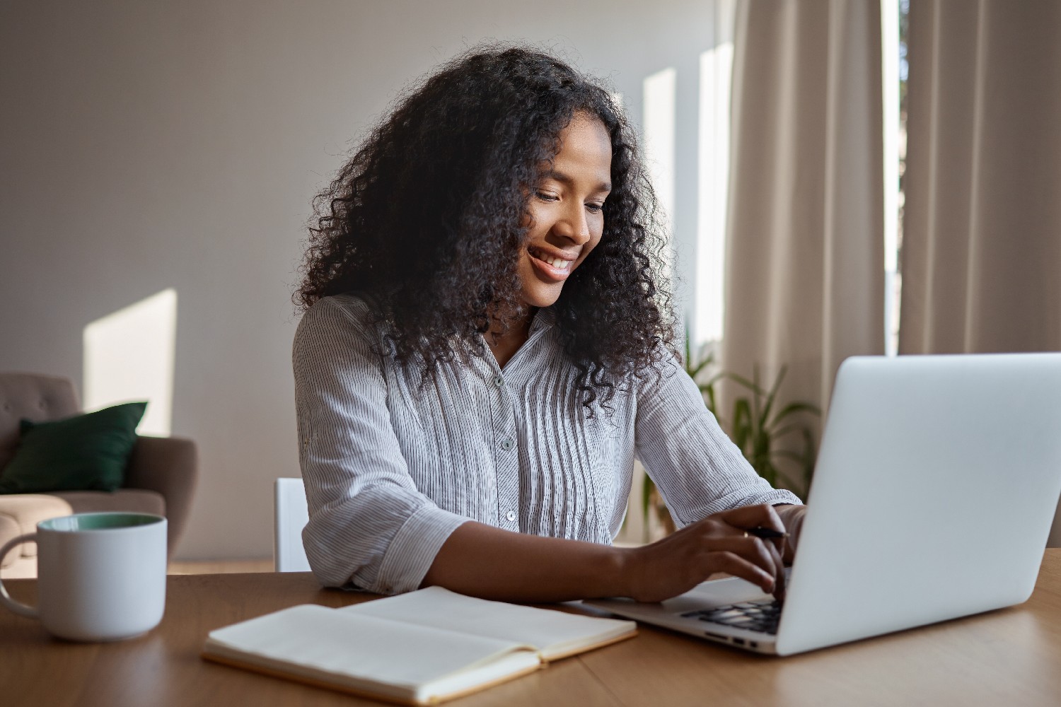 attractive-positive-young-afro-american-woman-freelancer-working-remotely-keyboarding-generic-laptop-sitting-home-with-copybook-mug-table-typing-electronic-message-online-smiling
